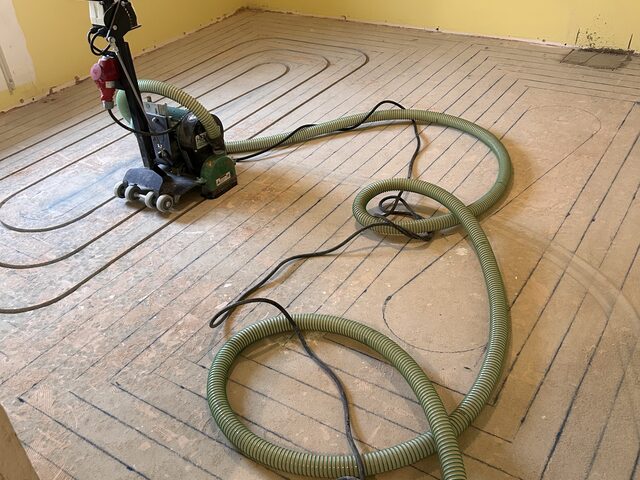 Zimmer mit gelber Wand während der Fräsarbeiten, Maschine und Absaugschlauch auf dem vorbereiteten Boden
